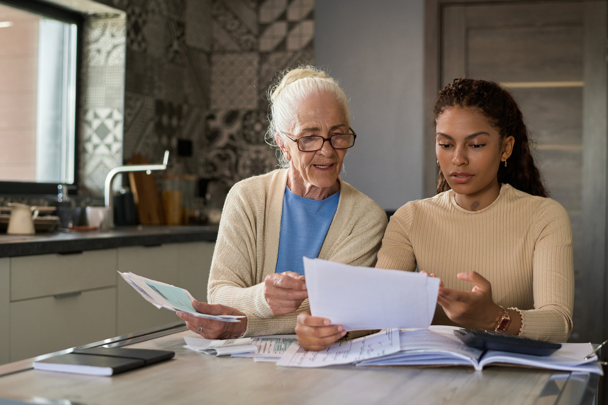 Grandmother and granddaughter sitting by table in the kitchen and discussing financial bills while one of them explaining information