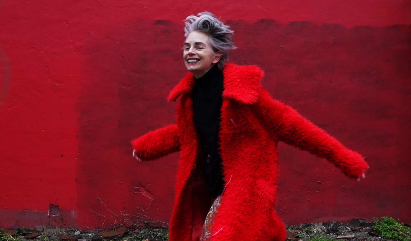 Woman with silver hair and red coat smiling in front of red wall