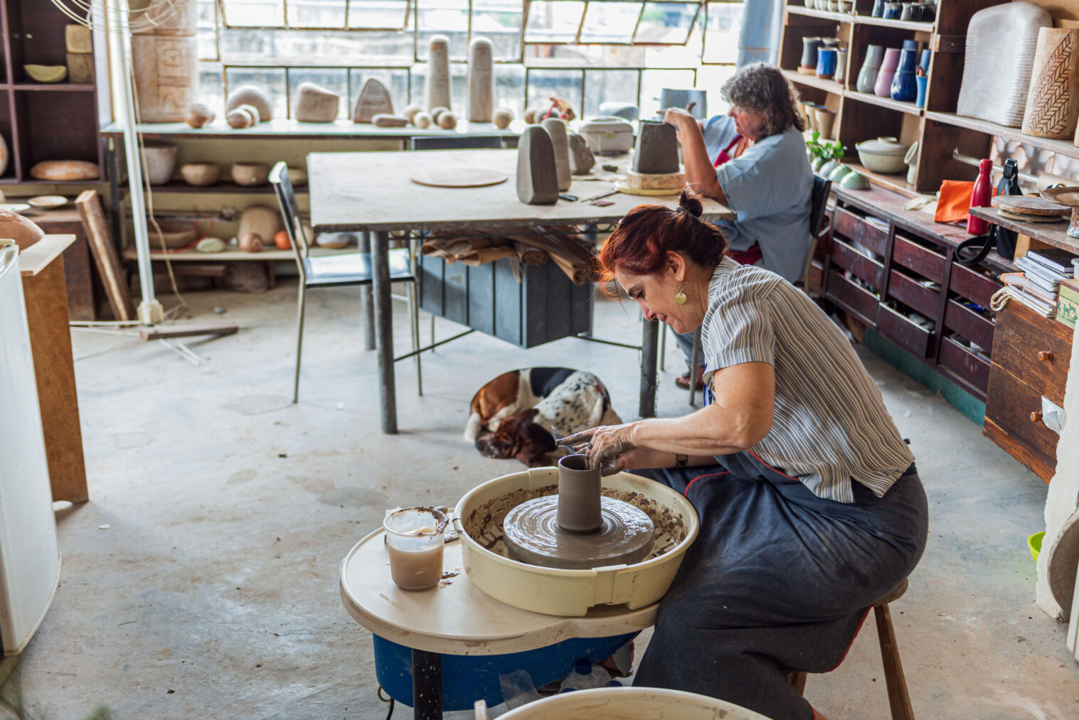 Older woman creating ceramic art with a pottery wheel. Another older woman is sculpting on a table in the back.